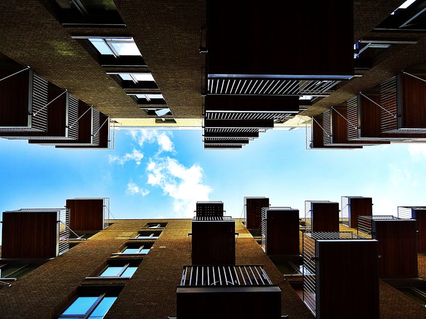 View looking up at a narrow courtyard between modern white and brick residential buildings in West Hampstead, with several black metal balconies attached to the white facade. The sky is visible above,