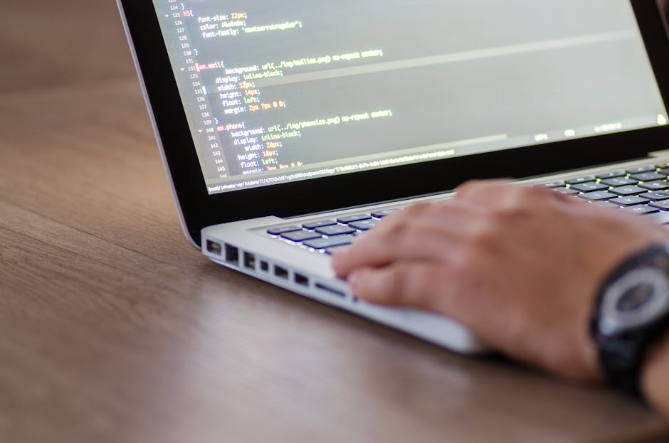 Close-up of a person's hand operating a laptop computer with visible lines of coding on the screen. The laptop is placed on a wooden surface, and the person's wrist is wearing a dark watch. The scene