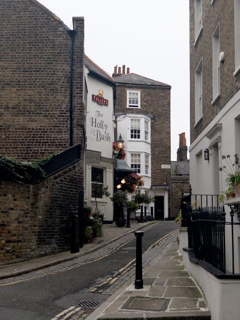 A narrow, sloped street in West Hampstead with brick and stone buildings on either side. On the left, a building with a sign reading 'The Holly Bush' features a white bay window and hanging flower bas