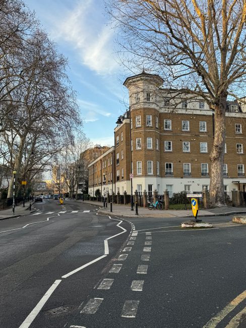 Photograph of a street scene in West Hampstead with a large multi-storey residential building featuring multiple windows, some with white window frames, and a rounded corner section at the top. Severa